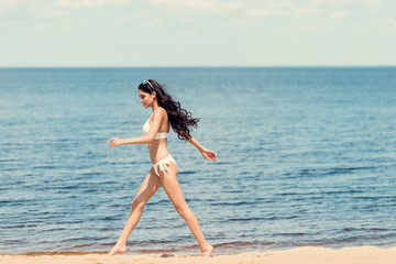 Naklejka premium happy young woman in white bikini running on beach near the sea
