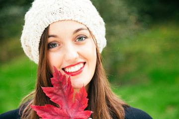 Beautiful young woman with woolen cap holding a red autumn leaf while smiling against a green background. Fresh skin and healthy smile.