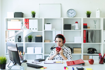 A young girl sits in headphones at a table in the office, holds a red cup in her hands and looks at the monitor.