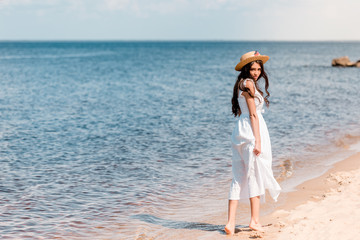 Fototapeta na wymiar young woman in straw hat and white dress walking on beach