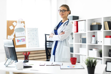 A young girl in a white coat is standing near a table in her office. A stethoscope hangs around her neck.