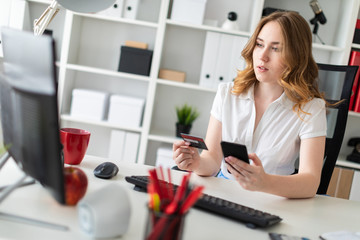 Beautiful young girl sits in the office, holds a bank card and phone in her hand.