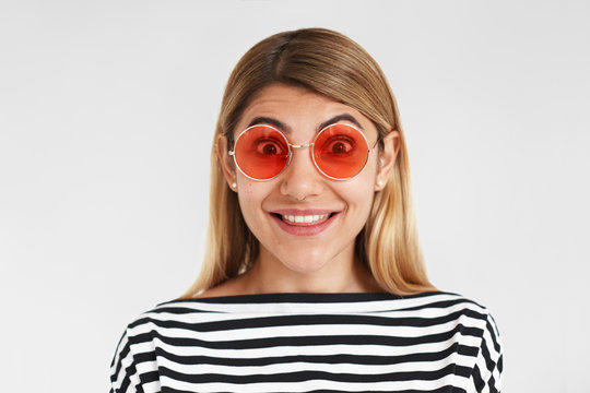 Isolated picture of funny emotional young Caucasian woman wearing trendy round sunglasses and striped top expressing excitement, joy, surprise or amazement, staring at camera, posing in studio