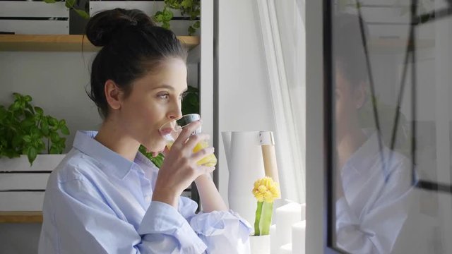 Young woman looking through window and drinking orange juice at home