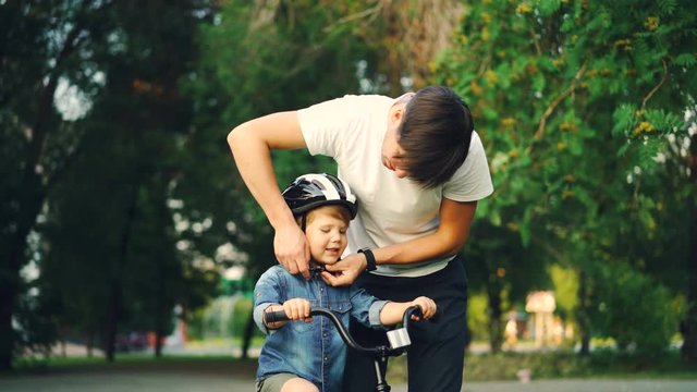 Caring father is putting safety helmet on his little son's head then teaching happy boy to ride bicycle while loving mother is watching them and smiling.