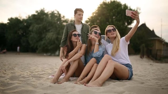 Shot of a group of young friends taking a selfie on the beach. Men and women taking photos sitting on a sand on warm summer evening.