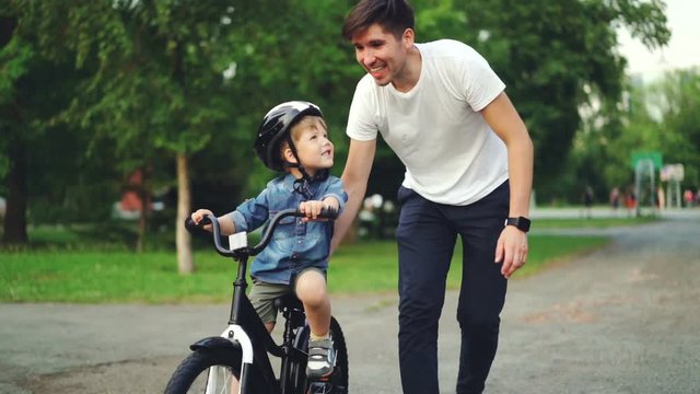 Slow motion of loving dad teaching his adorable son to ride bicycle in park holding bike and talking to child. Fatherhood, childhood and active lifestyle concept.