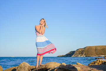 sexy blonde on the rocky coast of the sea, in a dress developing in the wind. She puts her hands together and pretends to be asleep.
