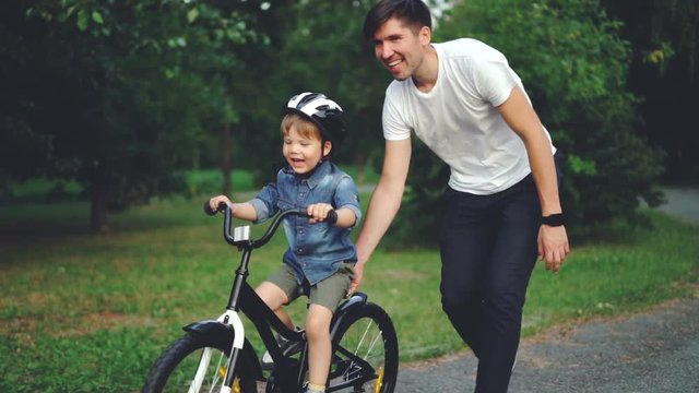 Slow motion of laughing child cycling in park with careful father who is teaching him to ride bicycle. Happy young family, fatherhood and childhood, active lifestyle concept.
