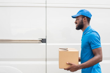 side view of smiling african american delivery man carrying cardboard box