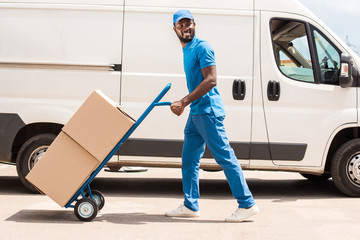 Fototapeta na wymiar side view of african american delivery man with cart and boxes