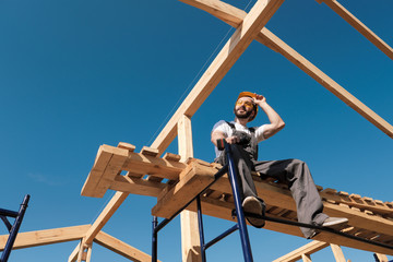 The man builder sits on the edge of the roof of the frame house, in a yellow helmet and gray...