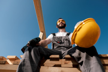 The man builder sits on the edge of the roof of the frame house, in a yellow helmet and gray overalls. The blue sky and clear sunny day.