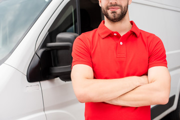 cropped shot of delivery man in red uniform with arms crossed standing at van