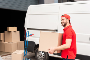 smiling delivery man in uniform with cardboard box standing near white van in street © LIGHTFIELD STUDIOS