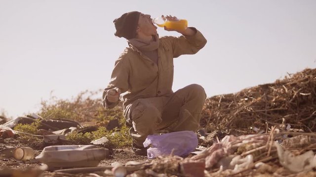 dirty homeless hungry man in a dump drinks the missing juice in the package with walking goes looking for food slow motion video. homeless dirty man roofless person looking for food in a dump. refugee