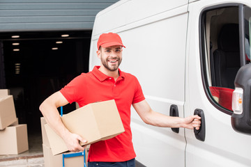 portrait of smiling delivery man in uniform with cardboard box standing near white van in street