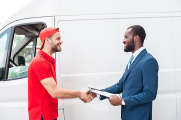 smiling delivery man and african american businessman shaking hands near van
