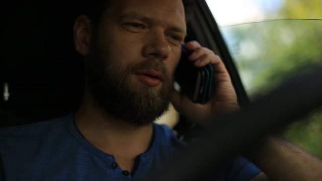 a bearded man sitting behind the wheel of a car and talking on the phone.