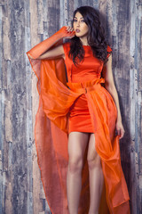 Fototapeta premium Young brunette woman in stylish orange silk dress posing on wooden background