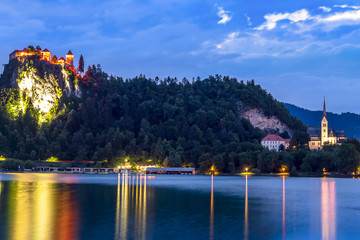 Fototapeta na wymiar The Bled Castle lighted during the blue hour overlooking the lake on a summer day. Slovenian castle on a cliff illuminated at night with water reflections