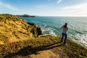 Woman walking along coastal trail taking smartphone photos