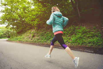 Young fitness blonde woman in headphones running at morning caucasian forest trail in sun light. side view from behind