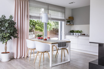Gray roman shades and a pink curtain on big, glass windows in a modern kitchen and dining room interior with a wooden table and white chairs - obrazy, fototapety, plakaty
