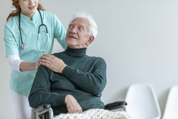 Doctor with stethoscope supporting disabled senior man in the wheelchair in a hospital