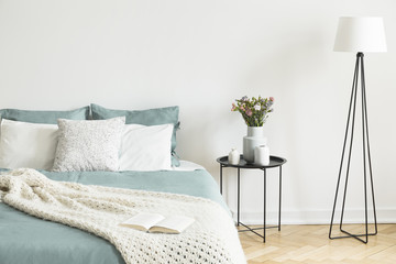 Real photo of bright bedroom interior with fresh flowers on bedside table, metal lamp and open book placed on bed with pastel bedding and knit blanket