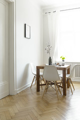 Wooden dining table with flowers in vase, two mugs and four chairs in real photo of bright room interior with window with drapes