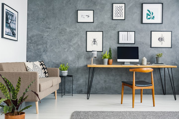 Brown couch next to wooden desk and chair in grey freelancer's interior with posters. Real photo