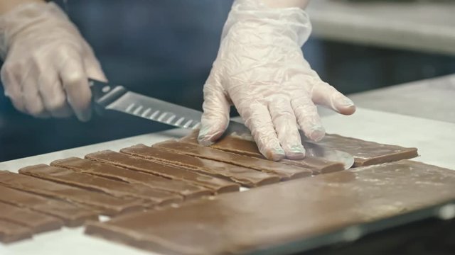 Close-up shot of female confectioner in gloves and apron cutting fudge with knife on paper