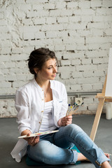 Fototapeta na wymiar Beautiful brunette girl in white shirt and jeans, sitting at work place and creating, drawing picture with oil colorful paints. Pretty woman, female artist holding brush and palette. Concept of art.