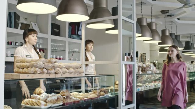 Young beautiful woman walking along glass display showcase in bakery and greeting friendly saleswoman