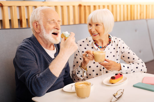 Yummy Dessert. Happy Senior Woman Sipping Coffee And Senior Man Biting Eclair