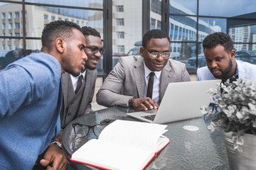 Group of happy diverse male business afro - american people team in formal gathered around laptop computer in bright office against the background of a glass building