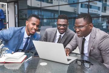 Group of happy diverse male business people afro american team in formal gathered around laptop computer in bright office against the background of a glass building