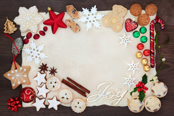 Christmas background border with joy sign including tree decorations, biscuits, cakes, chocolates and winter flora on parchment paper on rustic oak. Flat lay.