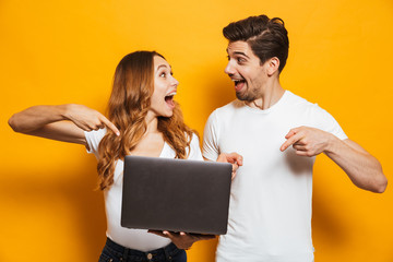 Portrait of excited happy man and woman holding and pointing fingers at black laptop, isolated over yellow background