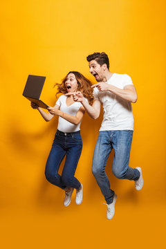Full length portrait of excited man and woman jumping and using black laptop, isolated over yellow background