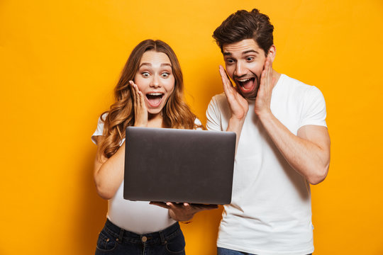 Portrait of excited man and woman screaming and holding black laptop, isolated over yellow background