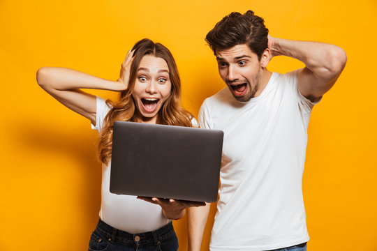 Portrait of excited couple man and woman screaming and grabbing head while holding black laptop, isolated over yellow background