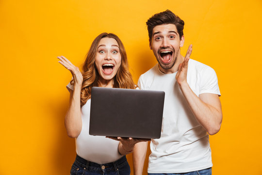 Portrait of excited man and woman screaming and rejoicing while holding black laptop, isolated over yellow background