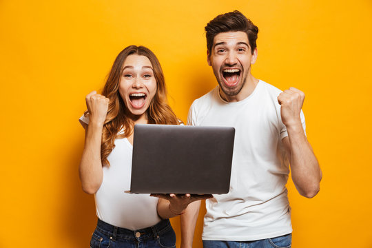 Portrait of excited man and woman screaming and clenching fists like winners or happy people while holding black laptop, isolated over yellow background