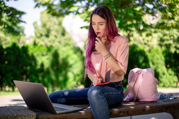 Woman writing in a notebook sitting on a wooden bench in the park. Girl working outdoors on portable computer, copy space.