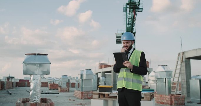 Young businessman in a suit with safety equipment on the rooftop of building speaking using a radio with some workers.