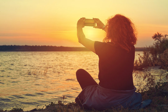 A young woman sits on the shore of a beautiful lake and takes a picture of a beautiful sunset on a smartphone. Family holiday in the countryside in nature. Panorama of a small lake. Orange sunshine