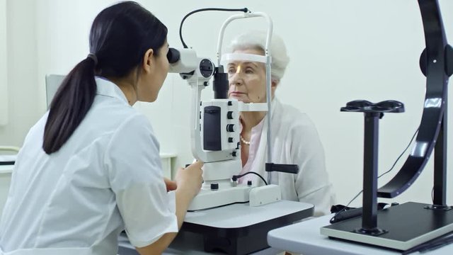 Panning shot of female optometrist examining eye of elderly woman with slit lamp in eye care clinic