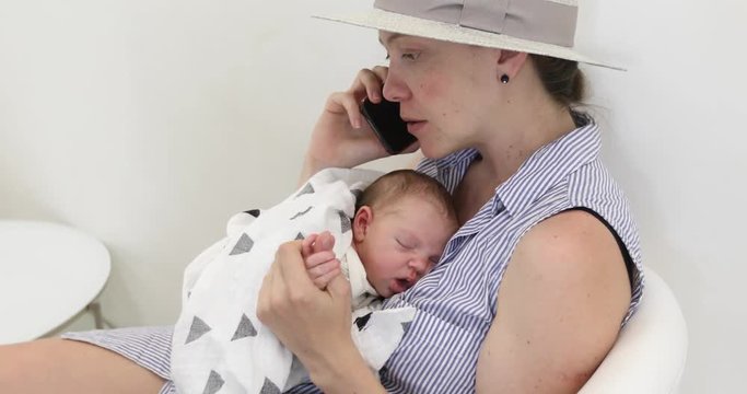Young mother in a gray straw hat speaks on the phone and holds a newborn baby in a white interior
